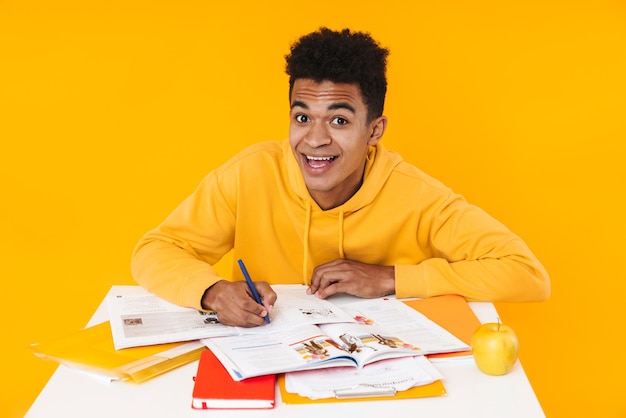 Happy african teenager boy studying while sitting at the desk and writing in textbook isolated on yellow