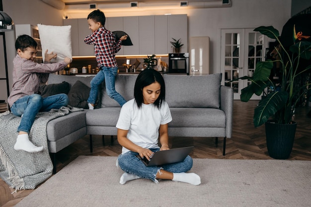 Happy african young mom working at home with laptop and adapting to active noisy sons fighting pillo