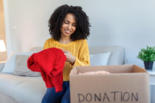 Happy African young woman sit on couch stuck clothes in donation box at home caring biracial female volunteer put apparel in carton package donate to needy people reuse recycle concept