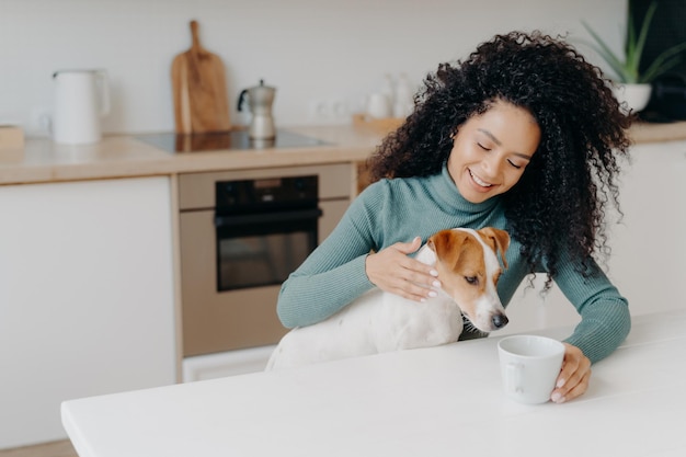 Happy Afro African woman with curly hairstyle treats dog in kitchen pose at white table with mug of drink enjoy domestic atmosphere have breakfast together People animals home concept