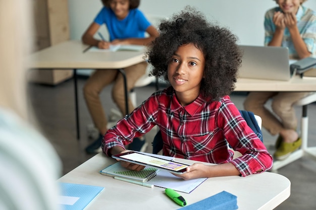 Happy Afro American schoolgirl looking at teacher holding tablet in classroom