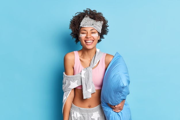 Happy Afro American woman smiles with teeth enjoys new day awakes in good mood had enough sleep wears comfortable pajama holds pillow appies patches under eyes stands indoor