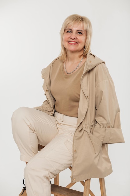 재킷을 입은 패션 베이지색 옷을 입은 행복한 나이의 여성은 흰색 배경에 있는 의자에 앉아 있습니다.