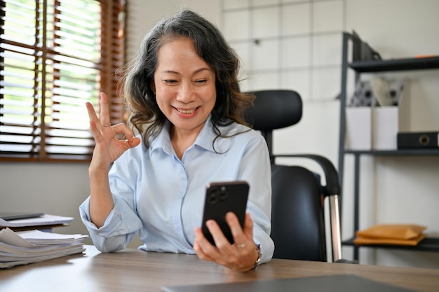 Happy aged Asian businesswoman making a video call through her smartphone making Okay hand sign