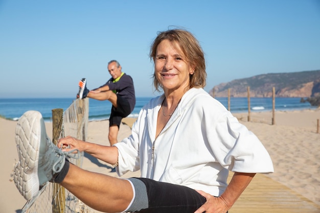 Happy aged couple training outdoors to keep fit. Smiling woman and man behind her spending time near sea doing sports and stretching legs. Sport activity and healthy lifestyle of aged people concept