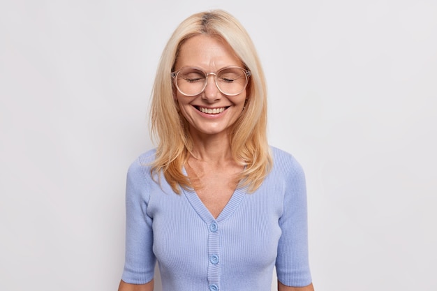 Happy aged female teacher glad to get congratulations with knowledge day glad to meet with pupils at school smiles joyfully keeps eyes closed wears big transparent gasses blue jumper poses indoor