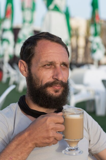 Happy american guy enjoying delicious cup of coffee. Portrait of handsome bearded man drinking hot espresso coffee with milk looks into the distance In sunset lighting. Man Holding A Coffee Mug.