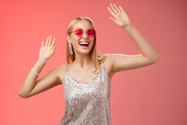 Happy amused carefree blond woman go wild dance-floor dancing having fun yelling yeah closed eyes waving hands moving rhythm music joyfully party in silver stylish dress sunglasses, red background.