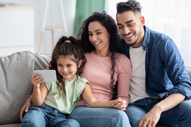 Счастливая арабская семья с маленькой дочерью, смотрящей видео на смартфоне дома, веселые ближневосточные родители и ребенок женского пола, расслабляющий с мобильным телефоном на диване в гостиной, крупным планом