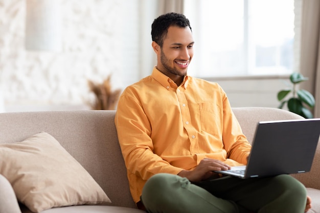 Happy arab man using laptop sitting on sofa at home