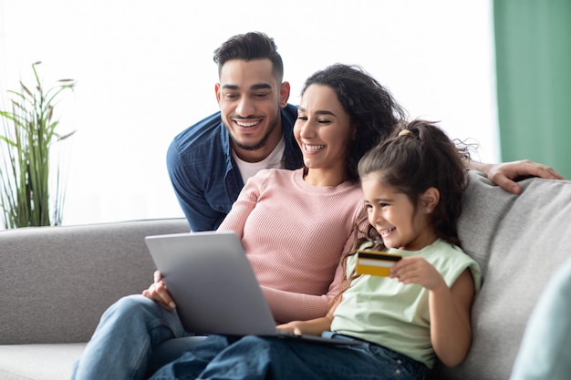 Photo happy arabic family using digital tablet and credit card for online shopping, relaxing on couch at home, young parents and their little daughter making purchases from internet together, free space