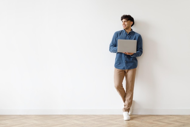 Happy arabic freelancer man holding laptop looking aside standing indoor