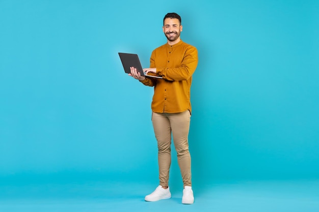 Happy arabic guy in freelancer daily life using modern laptop working online, blue studio background