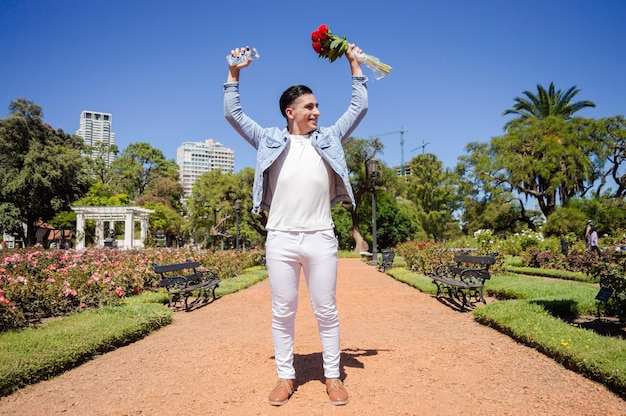 バラと木の美しい庭のある広場の真ん中に立っている幸せなアルゼンチンの白人男性は、花の花束と興奮して祝っている彼の携帯電話で手を上げています