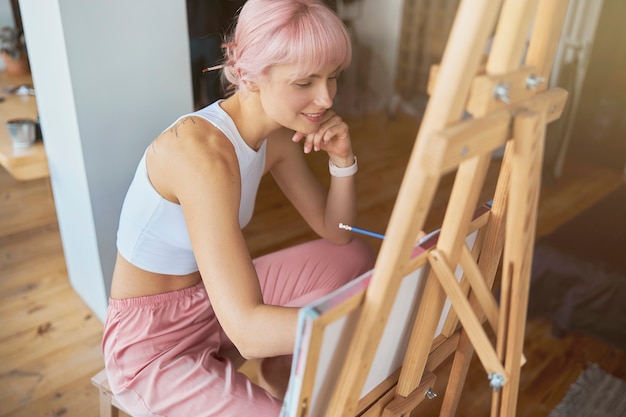 幸せな芸術家は木製のイーゼルに座って鉛筆で絵を描く
