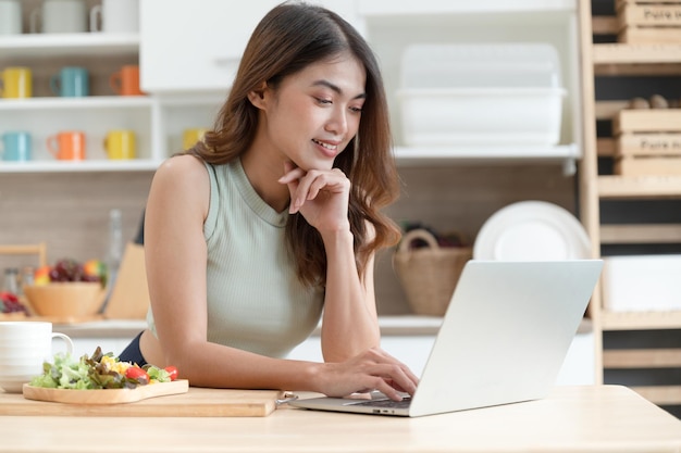 Foto la donna felice dell'asia che mangia insalata con vede il taccuino del computer nella stanza della cucina