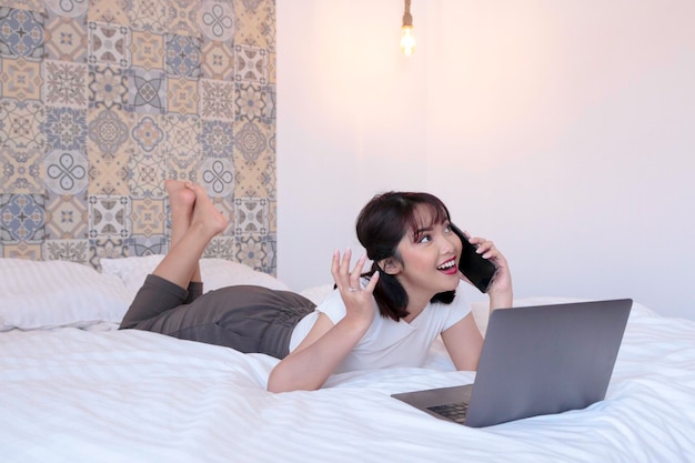 Happy asian beautiful girl work the laptop and make call in the bed Work from home concept