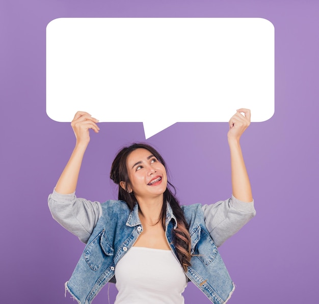 Happy Asian beautiful young woman smiling excited wear denims hold empty speech bubble sign, Portrait female posing show up for your idea looking at bubble, studio shot isolated on purple background