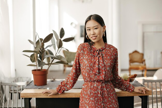 Happy Asian brunette woman in red stylish floral dress smiles looks into camera and leans on wooden table at kitchen