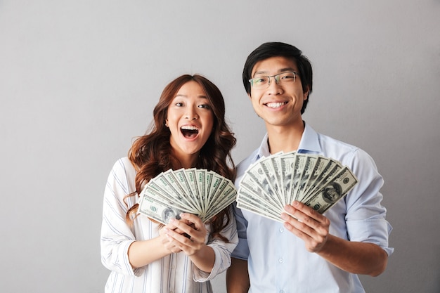 Happy asian business couple standing isolated, holding money banknotes