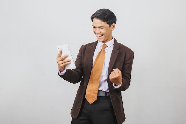 Счастливый азиатский бизнесмен в костюме и галстуке, держащий свой смартфон, празднует победный жест