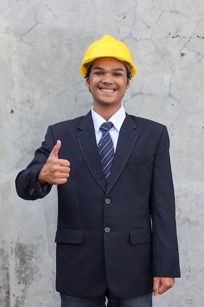 Happy Asian businessman in suit wearing safety yellow helmet smiling and give thumb up to the camera