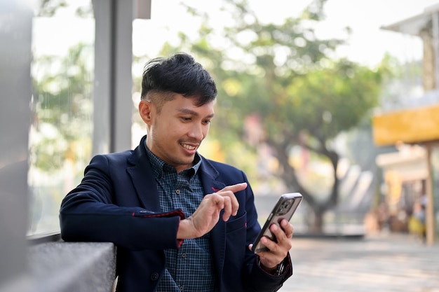 Happy Asian businessman using his smartphone while waiting for a bus or taxi in the city