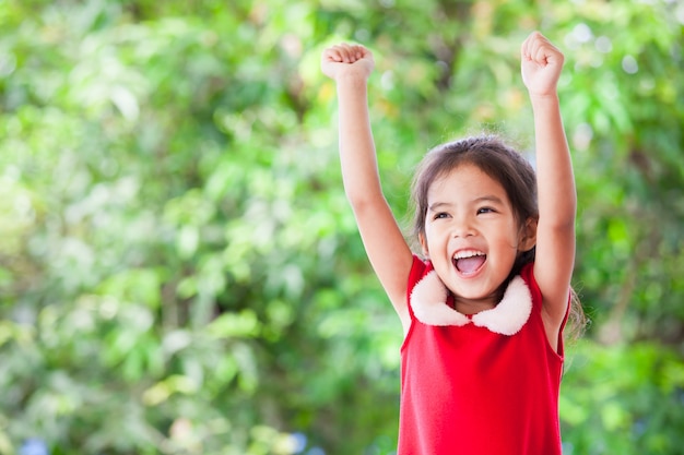 Happy asian child girl in christmas dress raising her hands and shouting with cheerful on green nature background