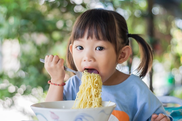 幸せなアジアの子供の女の子は一人でいくつかの麺を食べることを楽しんでいます。