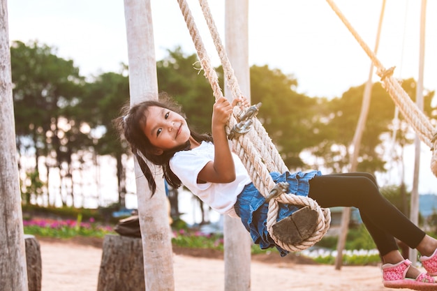 Happy asian child girl having fun to play on wooden swings in playground with beautiful nature 