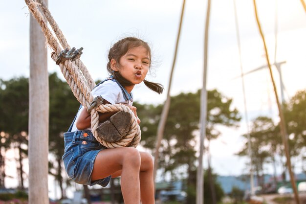 Happy asian child girl having fun to play on wooden swings in playground with beautiful nature 