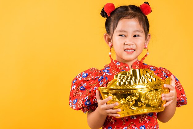大きな金のインゴットを保持している赤いチャイナドレスを着て幸せなアジアの中国の少女の笑顔