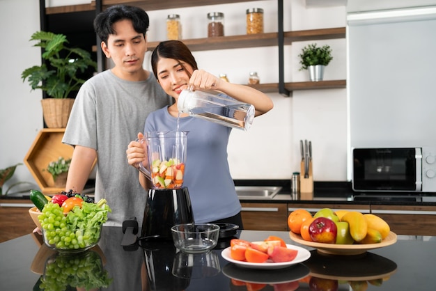幸せなアジアのカップルは、キッチン カウンターで健康的なビーガン スムージーを作るためのブレンダー マシンで新鮮な水をすすぐことを楽しんでいます 自宅で一緒にビーガン スムージーを作るカップル