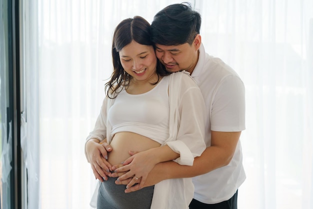 아기 를 기대 하고 있는 행복 한 아시아인 부부 가 집 의 창문 에 함께 서 있다