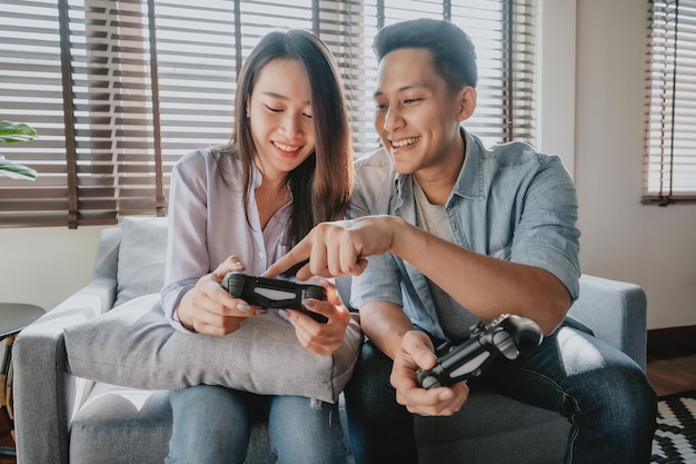 Happy asian couple having fun playing video game, man teaching\
woman how to play game at home