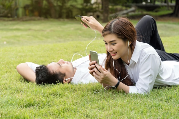 公園、恋人、ライフスタイルのコンセプトでスマートフォンのカメラを使って歌を聞いている幸せなアジアのカップル