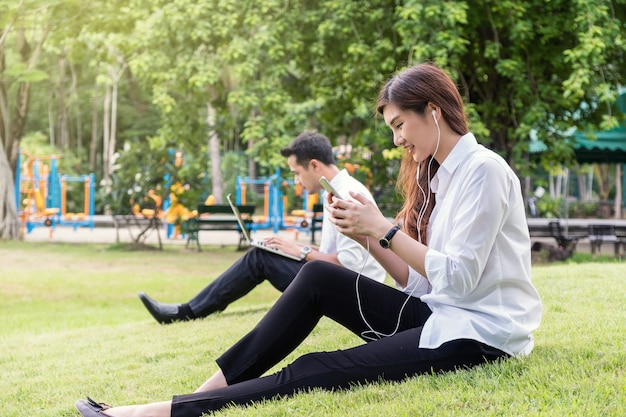 Happy asian couple listening the song via smart phone camera and using the laptop in the park, lover