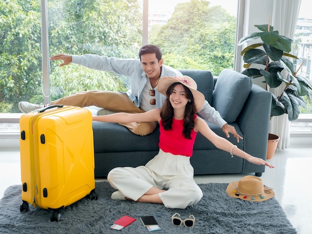 Happy asian couple lover ready to trip young woman and man in\
colorful casual making gesturing airplane wings smiling together\
sit on floor with passport in living room travel on summer\
holiday