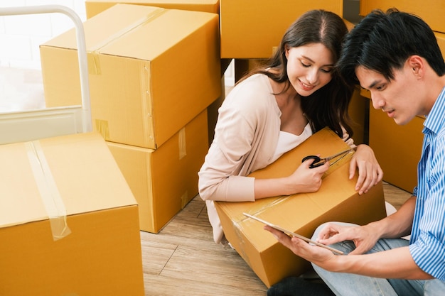 Счастливая азиатская пара переезжает в новый дом Женщина распаковывает картонные коробки из старого дома, в то время как мужчина делает покупки в Интернете с помощью цифрового планшета