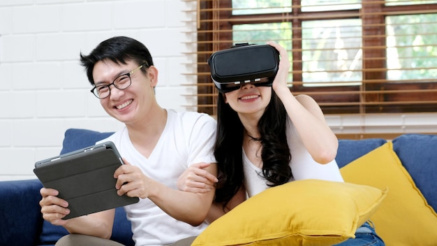 Happy asian couple play virtual reality simulation game while sitting on sofa at home living room, People and technology