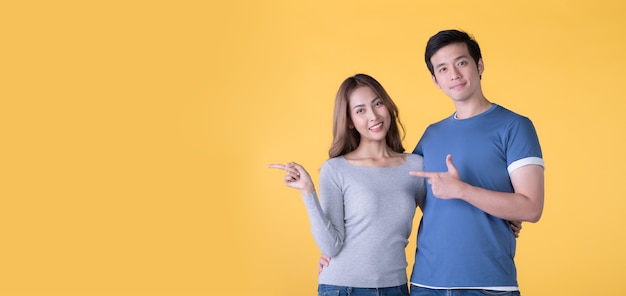 Happy Asian couple pointing fingers at empty copy space for text over yellow background