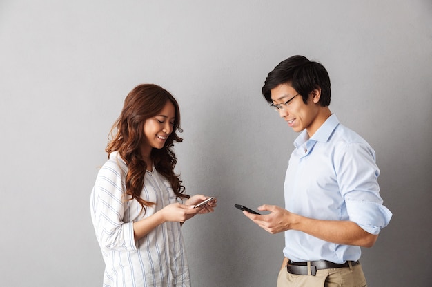 Happy asian couple standing isolated, using mobile phones