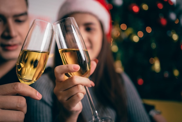 Happy Asian couple toasting champagne together sitting on the sofa in the evening with a Christmas tree and lights in the background Boyfriend and girlfriend at Christmas eve night