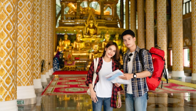 Happy Asian couple tourist backpackers holding paper map while traveling in beautiful Thai temple on holidays in Thailand, 