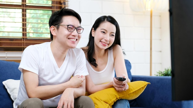 Happy asian couple watching television show while sitting on safa at home living room