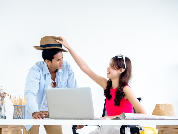 Happy Asian couple young woman put hat on man head with smile while using laptop computer on desk together for a trip information in home office ready to travel happy summer holiday concept