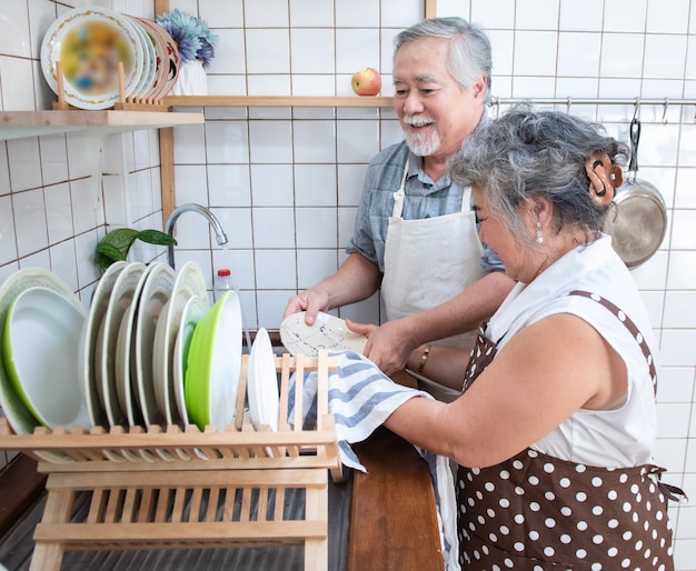 自宅の台所で自宅の流しで皿を洗って幸せなアジアの年配のシニアカップル。