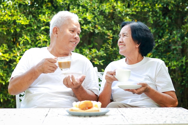 幸せなアジアの老夫婦が家の庭で朝一緒に座ってコーヒーを飲む 退職後の生活をお互いに世話する家族のコンセプト