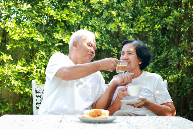 幸せなアジアの老夫婦が家の庭で朝一緒に座ってコーヒーを飲む 退職後の生活をお互いに世話する家族のコンセプト