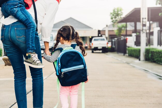 写真 幸せなアジアの家族の子供の男の子のピギーバックの母親と歩く歩道は屋外で子供の女の子を手で握り、母親は子供を学校に送り、学期のコンセプトの学校初日に戻る